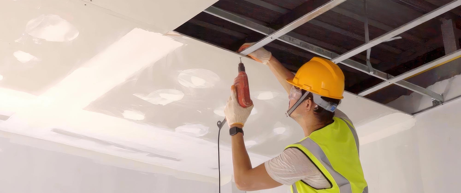 Construction worker in safety clothing and work gloves fastening the drywall ceiling to the metal frame using a drill